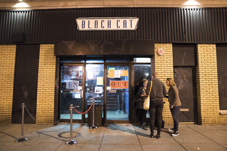 People stand outside the Black Cat nightclub under moody lighting on 14th Street, waiting to enter the venue.