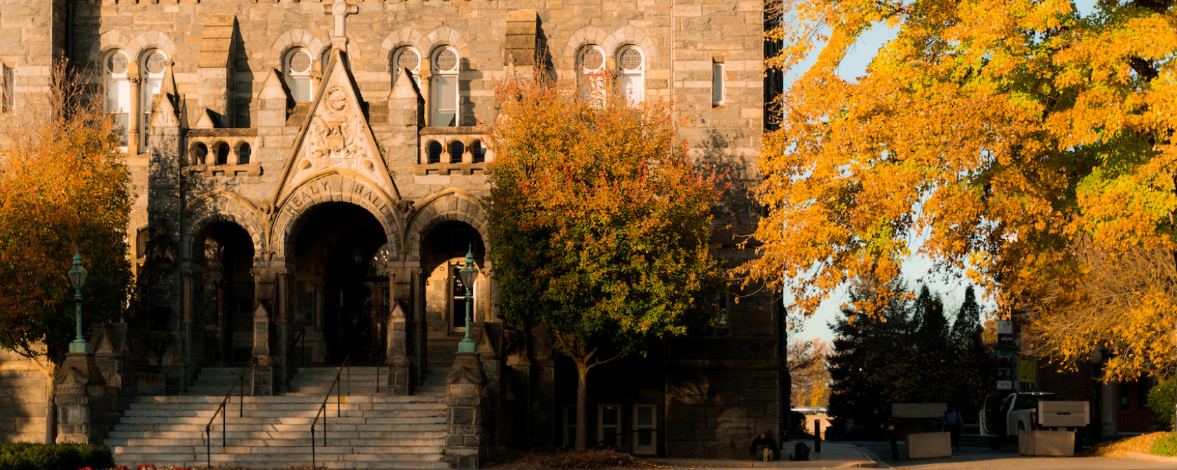 The historic Healy Hall at Georgetown University bathed in golden autumn sunlight, framed by vibrant orange and yellow foliage and a clear blue sky.