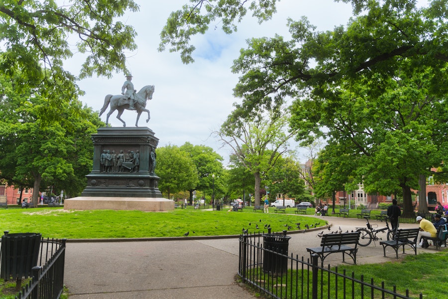 A wide view of Logan Circle Park featuring a large equestrian statue surrounded by leafy trees and benches on a sunny day.