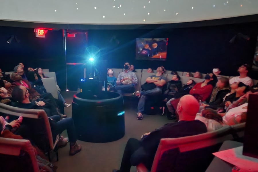 A group of children and adults gaze up at a starry projection during a show inside a darkened planetarium.