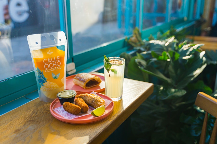Cuban pastries, croquettes, and lime cocktail with a Calle Ocho pouch at Colada Shop by a sunny window.