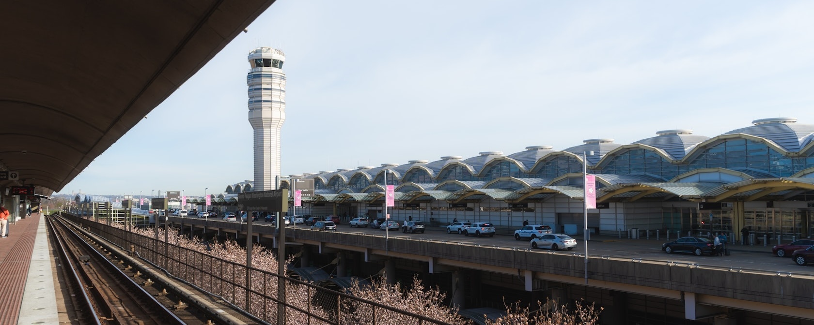 View of the exterior of Ronald Reagan Washington National Airport with the air traffic control tower and Metro tracks in the foreground on a clear day.