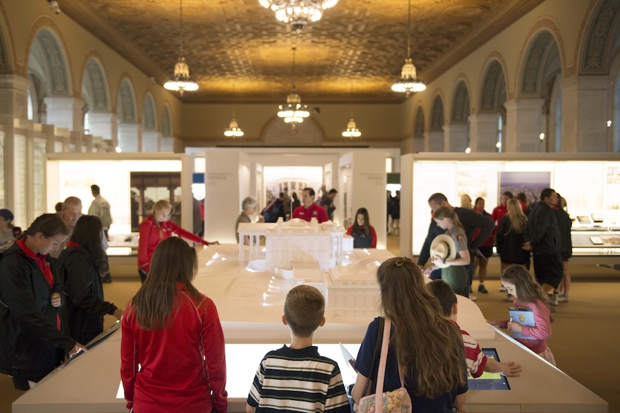 Visitors of all ages explore a scale model of the White House inside a grand exhibition hall.