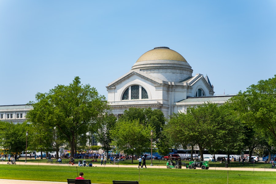 The Smithsonian National Museum of Natural History with its iconic dome, surrounded by trees and visitors walking along the National Mall.