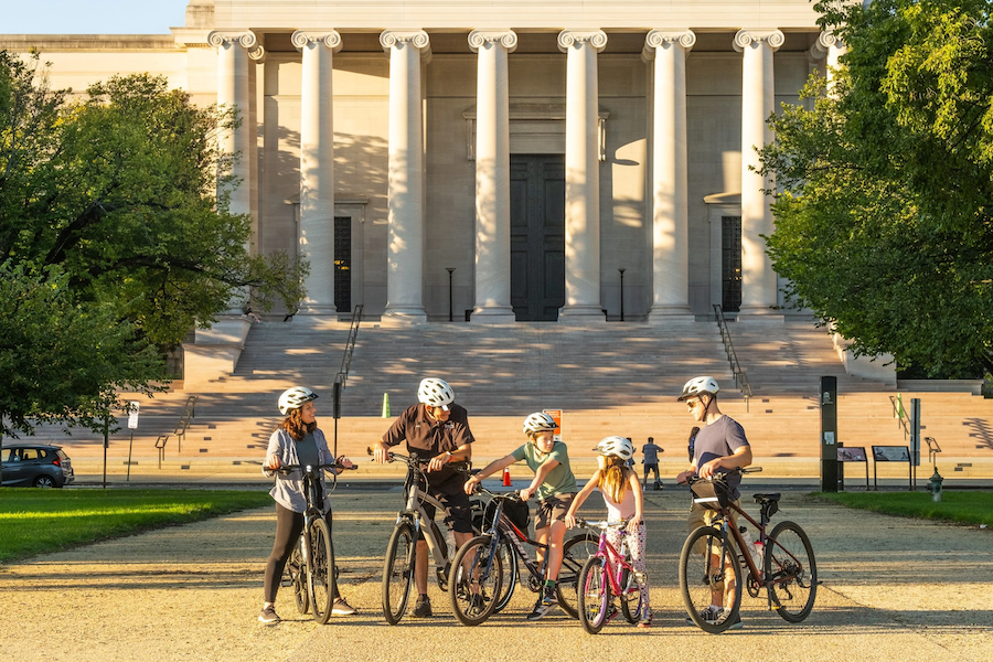A group of people with bicycles, including two children, stop in front of the National Gallery of Art in Washington, DC, during a guided bike tour.