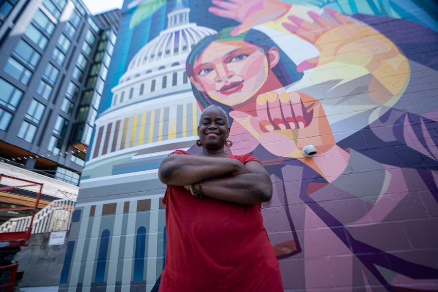 A person crosses their arms and smiles for a portrait in front of a colorful mural that depicts someone signing with the U.S. Capitol in the background. 
