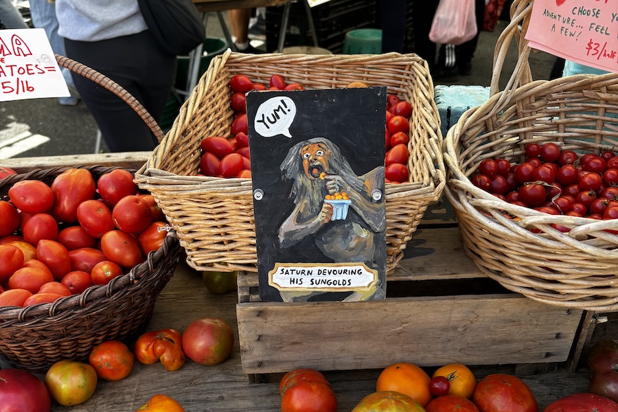 Baskets of tomatoes sit on display at a farmers market with a humorous sign reading “Saturn Devouring His Sungolds.”