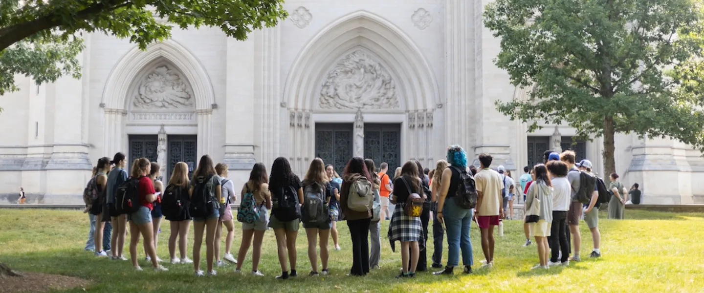 A group of people on a walking tour outside the Washington National Cathedral, standing on the lawn near the ornate stone entrance.