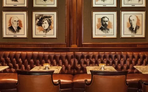 A cozy seating area featuring tufted leather banquettes, small marble tables, and portraits of notable historical figures adorning the walls in a warm wooden setting.