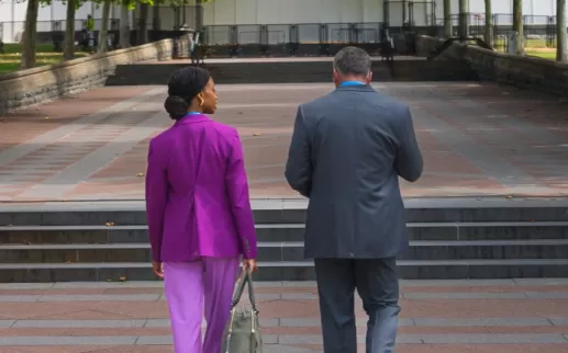 Two people walking on a brick sidewalk surrounded by green trees
