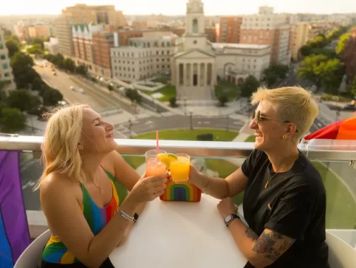 A couple enjoying a cocktail on a rooftop with a view of historic city buildings below. 