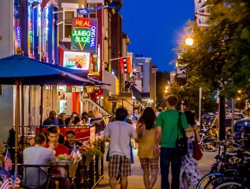 People walk down a street lit up with neon signs and busy restaurants and bars. 