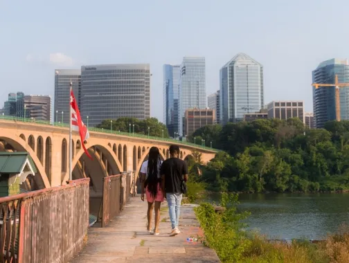 A couple walks along the Georgetown riverfront with a bridge and building of Arlington visible on the other side of the river. 