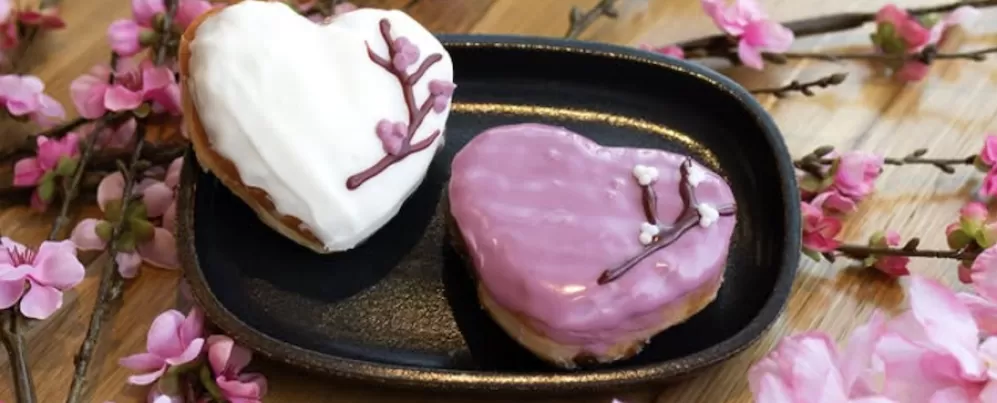 Two heart-shaped donuts decorated with cherry blossom branches sit on a table staged with cherry blossoms. 